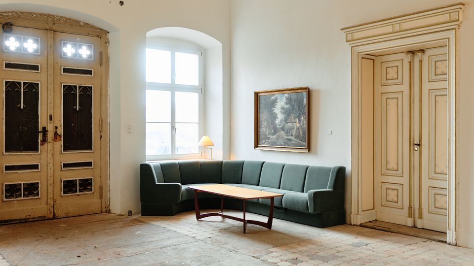 Heller Innenraum mit hohen weißen Wänden, großen Flügeltüren und einem hohen Fenster in einer Nische. In der Ecke steht ein L-förmiges grünes Sofa mit niedrigem Couchtisch; an der Wand hängt ein gerahmtes Bild, der Boden ist teils mit alten Fliesen ausgelegt.