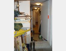 Ein Lager- oder Kellerraum mit offenen Regalen voller Werkzeuge und Baumaterialien auf der linken Seite. Im Hintergrund ist ein beleuchteter Flur mit einer offenen Tür zu sehen; Gummistiefel stehen auf dem Boden vor dem Eingang.