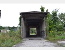 Verlassene Betonstruktur mit einem überdachten Durchgang, umgeben von überwuchernder Vegetation. Die Wände des Gebäudes sind mit Graffiti bedeckt, und im Hintergrund sind Bäume und Büsche sichtbar.