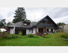 Einfamilienhaus mit Satteldach, weißer Fassade und schwarzen Schindeln auf dem Dach. Vor dem Haus liegt eine begrünte Rasenfläche, seitlich sind Gartenmöbel und ein Sonnenschirm erkennbar, im Hintergrund stehen Bäume.