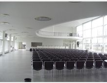 Großer, heller Veranstaltungssaal mit Reihen von schwarzen Stühlen, hohen Decken und einer großflächigen Fensterfront auf der rechten Seite. Im Hintergrund führt eine Treppe zu einer Galerie mit Geländer, und die Wände sind weiß gestrichen.