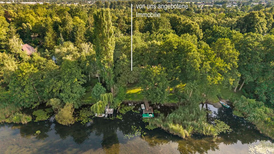 Luftaufnahme eines dicht bewaldeten Ufergrundstücks an einem See mit Schilf, Seerosen und mehreren kleinen Stegen mit Booten am Wasser. Im Hintergrund ist eine weitläufige Grünfläche mit vereinzelten Gebäuden am Horizont zu sehen, eine Markierung weist auf die angebotene Immobilie hin.