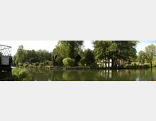 Panorama eines grünen Wasserkanals mit Bauminseln und einigen kleinen Gebäuden, darunter Holzhäuser mit Spitzdächern. Im Vordergrund ist ein Schild zu den Wasserständen sichtbar, die Umgebung ist von dichter Vegetation und Bäumen geprägt.