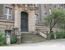 Fassade eines historischen Gebäudes mit steinernen Stufen, die zu einem verzierten Rundbogenportal mit schmiedeeisernem Doppeltor führen. Links und rechts des Eingangsbereichs befinden sich Fenster, die von detaillierten Steinornamenten umrahmt sind; vor dem Gebäude steht ein Baum.