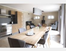 Helles Wohnzimmer mit offener Küche, einem großen Holztisch und gepolsterten Stühlen. Im Raum befinden sich außerdem ein Kaminofen, Sessel im Loungebereich und zwei kleine Fenster mit abgerundeten Kanten.