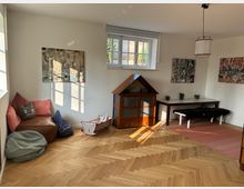 Ein heller Wohnraum mit Holzboden und weiß gestrichenen Wänden. Das Zimmer ist mit einer Leseecke mit Kissen und Sitzsack, einer kleinen Holzbank, einem Tisch, einem Puppenhaus und einer bemalten Kinderwiege eingerichtet.