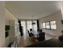 Esszimmer in einem modernen Haus mit großer Fensterfront und hellem Tageslicht. Der Raum zeigt einen langen Holztisch mit gepolsterten Stühlen, einen weißen Schrank an der linken Wand und helle Steinfliesen auf dem Boden; Dekorationen sind auf der Fensterbank platziert.