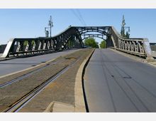 Metallbrücke mit geschwungener Konstruktion, über die eine zweispurige Straße sowie Straßenbahnschienen verlaufen. Im Hintergrund sind Bäume und Straßenelemente wie Laternen zu sehen; die Umgebung wirkt städtisch.