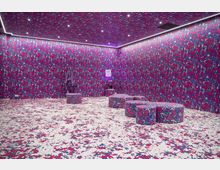 Ein Raum mit vollständig durchgehendem farbenfrohem Muster in Pink, Blau und Weiß auf Wänden, Boden und Möbeln. Im Raum befinden sich runde Hocker, eine beleuchtete Decke mit kleinen Einbauleuchten und schmale Lichtleisten entlang der Oberkanten der Wände.