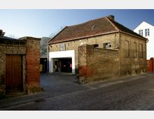 Backsteingebäude mit Satteldach und einigen abgenutzten Fenstern, daneben ein offenes Garagentor, hinter dem ein rotes Auto sichtbar ist. Im Vordergrund eine gepflasterte, kleine Straße und eine angrenzende Backsteinmauer.