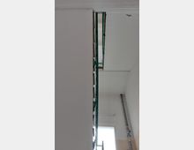 Treppenhaus mit weiß gestrichenen Wänden und Tageslicht, das durch ein rechteckiges Fenster einfällt. Ein begrenzender grüner Handlauf und eine vertikale Metallleitung sind sichtbar.