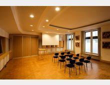 Ein moderner Seminarraum mit Holzboden und einer abgehängten, perforierten Decke mit eingebauten Leuchten. Der Raum enthält mehrere schwarze Stühle in Reihen vor einer Leinwand, daneben ein Holzpult; an den Wänden befinden sich Fenster mit Vorhängen und dekorative Holzelemente.