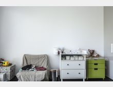 Raum mit hellem Interieur, einer grauen Polsterstuhlauflage sowie einer weißen Wickelkommode ausgestattet. Auf der Kommode liegen Wickelutensilien, daneben befindet sich ein grüner Rollcontainer und eine Tierfigur.