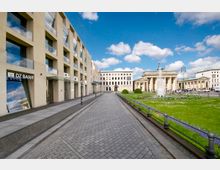 Straßenansicht mit modernen Gebäuden auf der linken Seite, darunter ein DZ Bank-Geschäft, und dem Brandenburger Tor im Hintergrund rechts. Im Vordergrund eine breite gepflasterte Gehwegstraße mit angrenzenden Grünflächen und einem Brunnen.