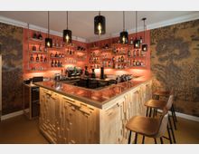 Moderner Innenraum einer Bar mit einer Theke aus Holz und einem glänzenden Tresen, umgeben von Regalen mit Flaschen und Gläsern. Die Wände sind in einem orangenen Farbton gestaltet und mit einem floralen Muster dekoriert, während drei Hängelampen mit sichtbaren Glühbirnen den Raum beleuchten.