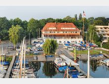 Luftaufnahme eines kleinen Hafens mit Segelbooten, Anlegestegen und einem großen Gebäude mit rotem Dach im Hintergrund. Umgeben von Bäumen und Rasenflächen, weht auf einem Mast die deutsche Flagge.