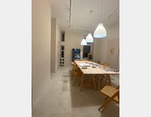 Langer, heller Atelierraum mit hohen weißen Wänden und Betonboden, in der Mitte ein großer Holztisch mit mehreren Holzstühlen und ausliegenden Mal- und Bastelmaterialien. Über dem Tisch hängen weiße Pendelleuchten, im Hintergrund stehen mehrere Gemälde an der Wand und bei einem Fenster.