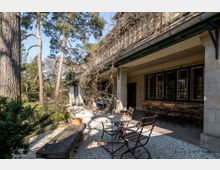 Steinveranda eines historischen Gebäudes mit Metalltischen und Holzstühlen auf einem mit Kies bedeckten Boden, umgeben von Bäumen und einem Garten. Die Fassade zeigt große Fenster mit Gitterelementen, und Kletterpflanzen ranken an der Veranda empor.