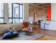 Wohnbereich in einem Loft mit sichtbaren Backsteinwänden, großen Fenstern mit blauen Rahmen und Holzboden. Im Vordergrund steht ein brauner Ledersessel auf einem gemusterten Teppich, während im Hintergrund eine offene Küche und ein Essbereich zu sehen sind.