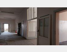 Ein leerer Flur mit weißen Wänden und Fliesenboden. Mehrere geschlossene und geöffnete Türen führen zu angrenzenden Räumen, einige mit Glasscheiben; im Hintergrund ist ein Fenster mit einem Heizkörper darunter erkennbar.