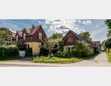 Eine ländliche Szene mit mehreren Wohnhäusern, die von Bäumen und Gärten umgeben sind. Die Gebäude haben gelbe Fassaden, rote Satteldächer und kleine Fenster mit Holzläden; im Vordergrund befindet sich eine gepflasterte Straße.