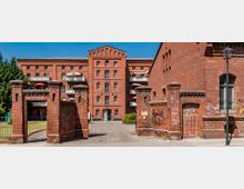 Backsteingebäude im historischen Industriearchitekturstil, umgebaut zu Wohnanlagen mit Balkonen. Im Vordergrund ein Torbogen aus roten Ziegelsteinen, umgeben von mit Graffiti bedeckten Mauern; rechts eine Laterne und links ein grüner Gartenbereich.