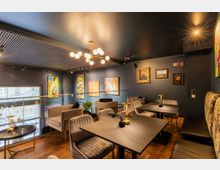 Ein modernes Café mit dunklen Wänden, eingerichtet mit eleganten Tischen, Polstersesseln und Wandgemälden. Große Fenster bieten Tageslicht, während diverse Kunstwerke die Wände schmücken und eine gemütliche Atmosphäre schaffen.