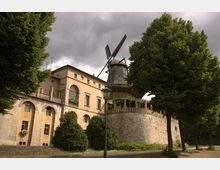 Historisches Gebäude mit gelblicher Fassade und Rundbogenfenstern, daran angebaut eine alte Windmühle mit dunklem, schindelgedecktem Dach. Umgeben von Bäumen und einem gepflasterten Rundgang auf einer steinernen Plattform.