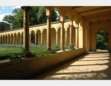 Eine überdachte Kolonnade mit gelblichen Rundbögen und Steinsäulen, umgeben von einem gepflegten Garten mit grünem Efeu und Büschen. Im Hintergrund sind Bäume und ein Teil eines Gebäudes sichtbar.