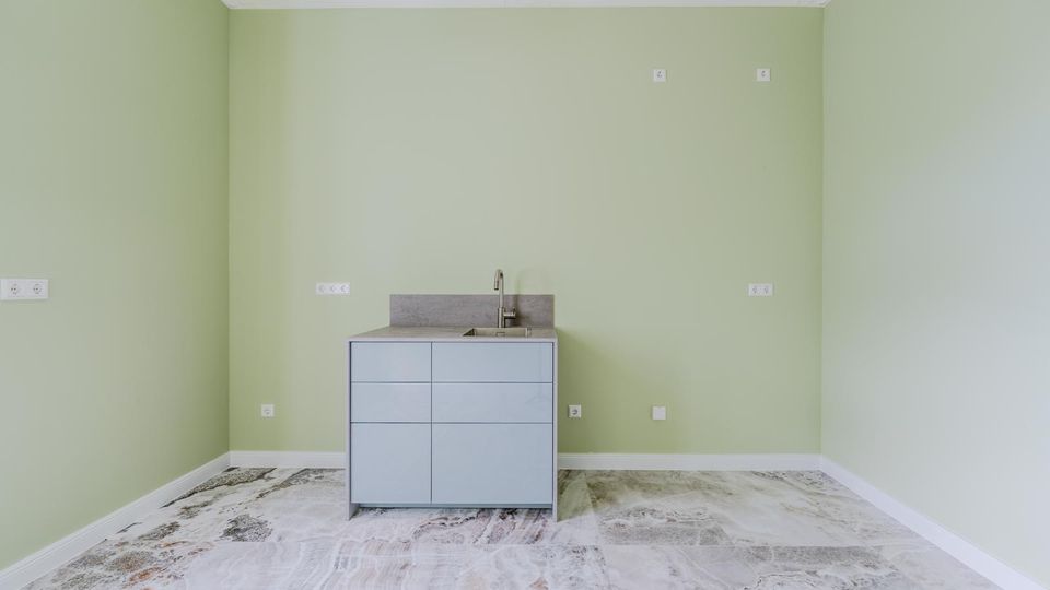 Leerer Küchenraum mit hellgrünen Wänden, Stuckdecke und marmoriertem Steinboden. In der Mitte steht ein grauer Unterschrank mit Spüle und Armatur; mehrere Steckdosen und Anschlüsse sind an der Wand verteilt.