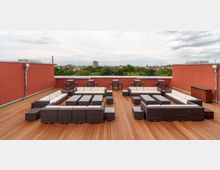 Dachterrasse mit Holzboden, flankiert von roten Wänden, ausgestattet mit modernen Sitzgruppen aus dunklem Rattan und hellen Polstern sowie Stehtischen im Hintergrund. Im Hintergrund ist eine grüne Baumlandschaft und eine städtische Skyline sichtbar.
