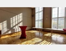 Heller Raum mit großen, bodentiefen Fenstern mit vertikalen Lamellenjalousien, die Licht- und Schattenmuster auf den hölzernen Parkettboden werfen. Im Raum stehen ein Stehtisch mit rotem Stoffbezug und ein langer, schmaler Tisch.