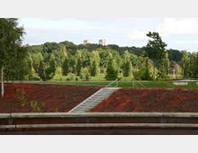 Parklandschaft mit einem bepflanzten Hügel, in dessen Mitte eine von Handläufen flankierte Treppe verläuft. Im Hintergrund befinden sich dichte grüne Bäume, und auf einem bewaldeten Hügel sind zwei Türme sichtbar.