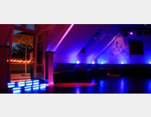 Ein schummrig beleuchteter Innenraum eines Nachtclubs mit blauer und violetter Beleuchtung. Links führen beleuchtete Stufen durch eine geöffnete Tür zu einer Terrasse mit Sonnenschirm, während rechts Sitzbereiche und eine Wand mit projiziertem Bild sichtbar sind.