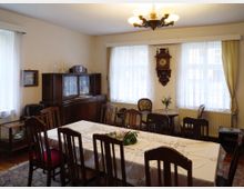 Wohnzimmer mit klassischem Mobiliar, darunter ein großer Esstisch mit Stühlen, eine hölzerne Kredenz mit Glastüren, ein Wanduhrgehäuse und ein kleiner Beistelltisch mit Blumen. Die Fenster sind mit weißen Vorhängen dekoriert, durch die Tageslicht einfällt.