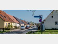 Straßenszene in einem Wohngebiet mit Einfamilienhäusern, die dicht an einer ruhigen Straße liegen; die Häuser haben rote Ziegeldächer, einige mit Solarpanelen. Ein Straßenschild mit der Aufschrift „Bammer Landstraße“ und ein Halteverbotsschild sind im Vordergrund sichtbar.
