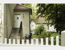 Eingang eines beige gestrichenen Hauses mit einer weißen Tür, die über eine kleine Steintreppe erreichbar ist. Im Vordergrund ein weißer Zaun, ein Fahrrad mit Kindersitz sowie umliegende grüne Büsche und Bäume; im Hintergrund ein Gebäude mit Ziegeldach.