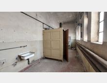 Verlassener Raum mit hohen Decken, abgenutzten Wänden und einem kleinen Waschbecken an der linken Wand. Auf der rechten Seite sind mehrere Fenster mit Gitterglas sowie alte Rohrleitungen zu sehen; im Hintergrund steht eine geöffnete Schranktür.