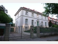 Weiß gestrichene Stadtvilla im klassischen Stil mit grüner Eingangstür und filigranem Balkongeländer, umgeben von einem Garten mit Blumenbeeten. Vor der Villa befindet sich ein schmiedeeiserner Zaun mit einem gepflasterten Eingangsbereich.