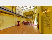 Großer, gelb gestrichener Veranstaltungsraum mit hohen Decken und großzügigen Fenstern, dekoriert mit Musterelementen an den Wänden. Im Raum steht ein U-förmiger Konferenztisch mit roten Stühlen, während ein Kronleuchter von der Decke hängt und eine Galerieebene mit Geländer den oberen Bereich überblickt.