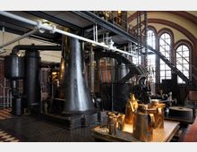 Historische Maschinenhalle mit einer großen schwarzen Dampfmaschine, Metallrohren und Zahnrädern, umgeben von roten Ziegelwänden mit hohen Rundbogenfenstern. Im Vordergrund stehen glänzende, goldfarbene Ölkanister auf einem Arbeitstisch.