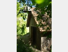 Holzhütte mit Schindeldach in einem bewaldeten Gebiet, teilweise von grünen Blättern umrahmt. Der Zugang führt über einen steinigen Pfad, und die Umgebung ist von dichtem Laub geprägt.