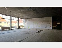 Leerer, verlassener Innenraum eines ehemaligen Gewerberaums mit großflächigen Fensterfronten zur Straße, abgenutztem Holzboden und dunkler Rasterdecke mit freihängenden Leitungen. An der Rückwand sind beschädigte Verkleidungen und Graffiti zu sehen, daneben ein offener Durchgang zu einem Nebenraum.