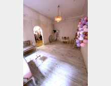 Ein leerer Raum mit hölzernem Dielenboden, einer cremefarbenen Wand mit sichtbaren Gebrauchsspuren, einem Kronleuchter an der Decke und einem Arrangement aus bunten Luftballons an der rechten Ecke. Im Hintergrund ist durch einen Rundbogen ein Bereich mit Kleidung auf Ständern sichtbar.