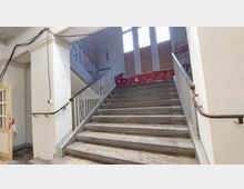 Ein Treppenhaus in einem verlassenen Gebäude mit breiter Steintreppe, Metallgeländern und Fenstern im Hintergrund, die mit Holzplatten abgedeckt sind. Wände und Geländer sind teilweise mit Graffiti und Abnutzungsspuren versehen.
