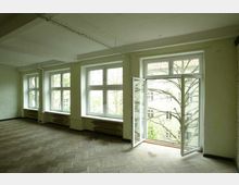 Ein leerer heller Raum mit großen Fenstern und einer geöffneten Doppeltür, die auf einen Balkon führt. Der Boden ist aus Fischgrätparkett, und durch die Fenster sind Bäume sowie ein gegenüberliegendes Gebäude sichtbar.