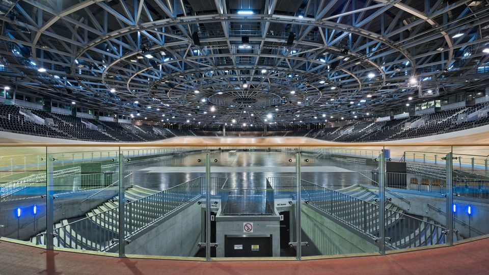 Innenansicht einer großen Radrennbahn-Halle mit ovaler Holzbahn, dunklen Tribünenreihen und einer zentralen Infield-Fläche. Über dem Innenraum spannt sich eine komplexe Stahlträger-Dachkonstruktion mit ringförmigen Beleuchtungselementen und vielen Scheinwerfern.