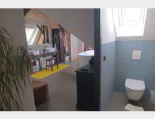 Modernes Badezimmer unter einem Dach mit Schrägen; links ein Waschtisch mit großem Spiegel und freiliegenden Backsteinwänden, rechts eine Toilette vor blau gefliester Wand und ein Dachfenster, im Hintergrund eine freistehende Badewanne.