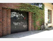 Backsteingebäude mit schwarzem Tor, das mit Graffiti besprüht ist. Links und rechts des Tors sind Kletterpflanzen an der Fassade, und daneben steht ein Fahrrad an der Wand gelehnt.