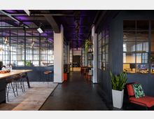 Moderner Büroraum mit industriellem Design, gekennzeichnet durch verglaste Trennwände mit schwarzen Metallrahmen, einem hohen Tisch mit Barhockern und einer offenen Decke mit sichtbarer Beleuchtung. Pflanzen, dekorative Regale und Sitzgelegenheiten ergänzen die Einrichtung entlang eines breiten Flurs.